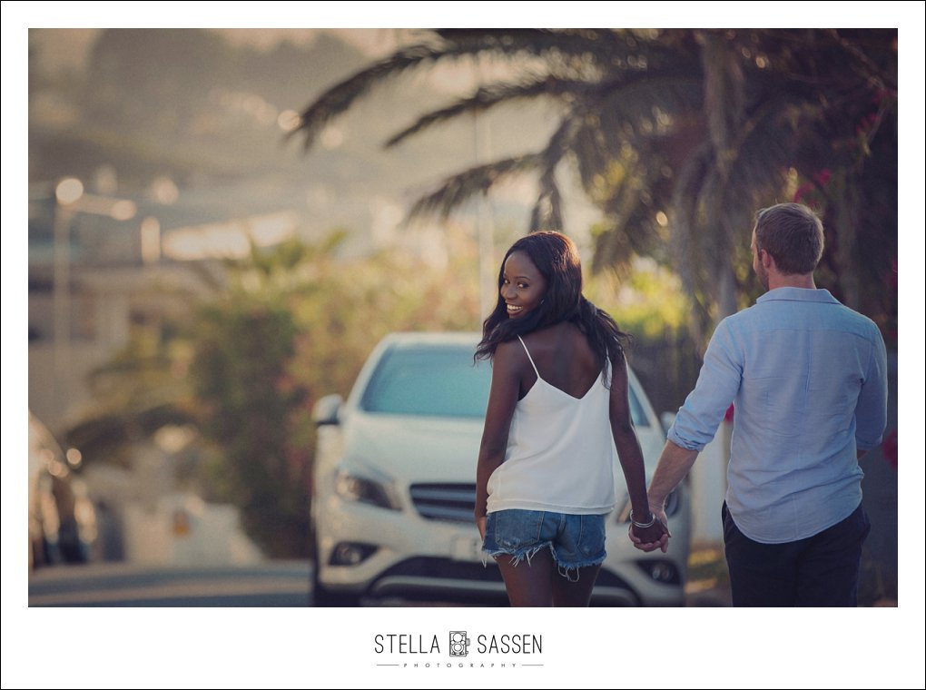 A couple walking hand in hand through the streets of Camps Bay at golden hour, the woman glancing back over her shoulder with a wide smile, palm trees and the warm Cape Town light behind them.