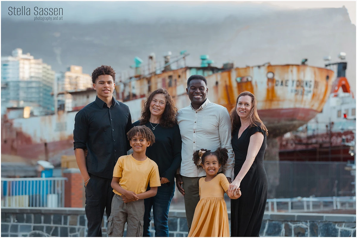 A family of six photographed at the V&A Waterfront in Cape Town at sunset, with the iconic rusted fishing vessel and Table Mountain visible in the background.