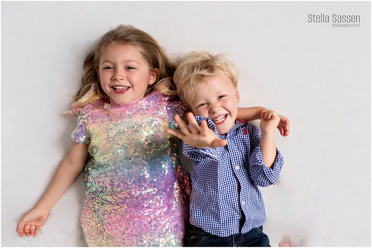 Two young siblings laughing and lying together on a white studio backdrop during a family photography session in Cape Town — a girl in a rainbow sequin dress and her brother in a checked shirt waving at the camera.