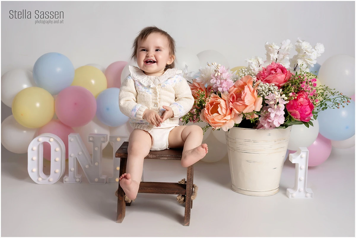 A one-year-old girl laughing during her cake smash birthday session in a Cape Town studio, seated on a wooden stool surrounded by pastel balloons, fresh flowers, and lit "ONE" and "1" props.