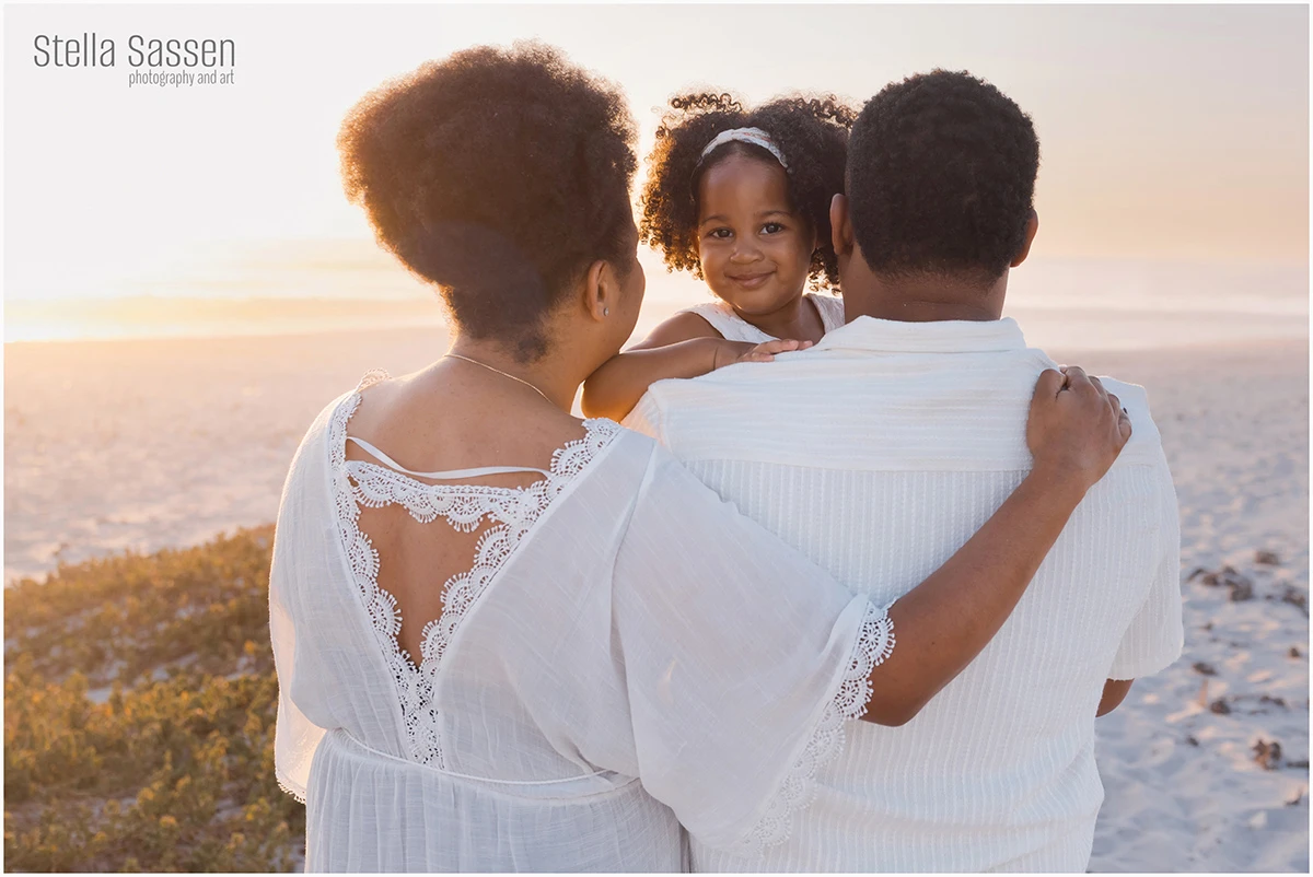 A family of three at golden hour on a Cape Town beach, parents facing away from the camera while their young daughter, perched on her father's back, smiles at the camera. All three are dressed in white.