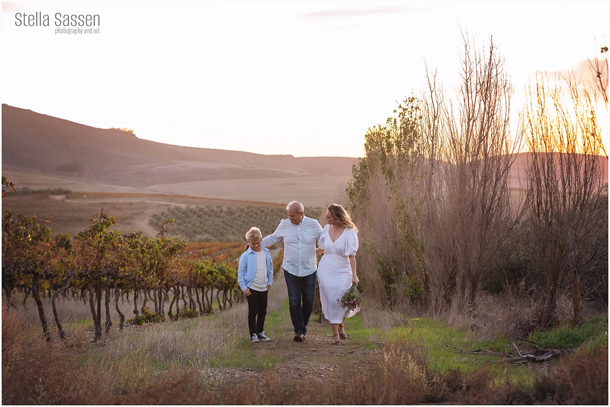 A family of three walking together through a Cape Winelands vineyard at sunset, autumn vines and rolling mountains behind them, mom carrying a wildflower bouquet.
