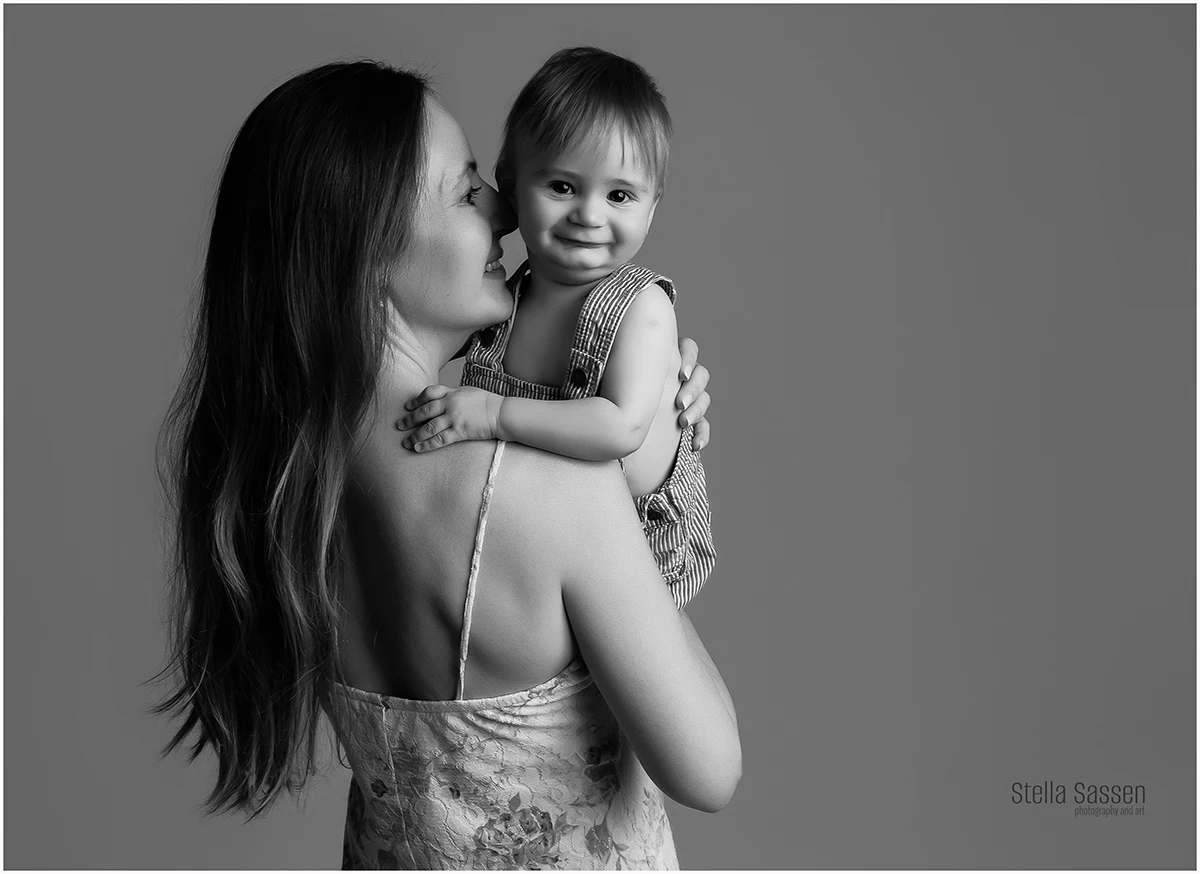 Black and white studio portrait of a mother kissing her baby boy on the cheek as he smiles at the camera, captured during a mommy and me sitter session in Cape Town.