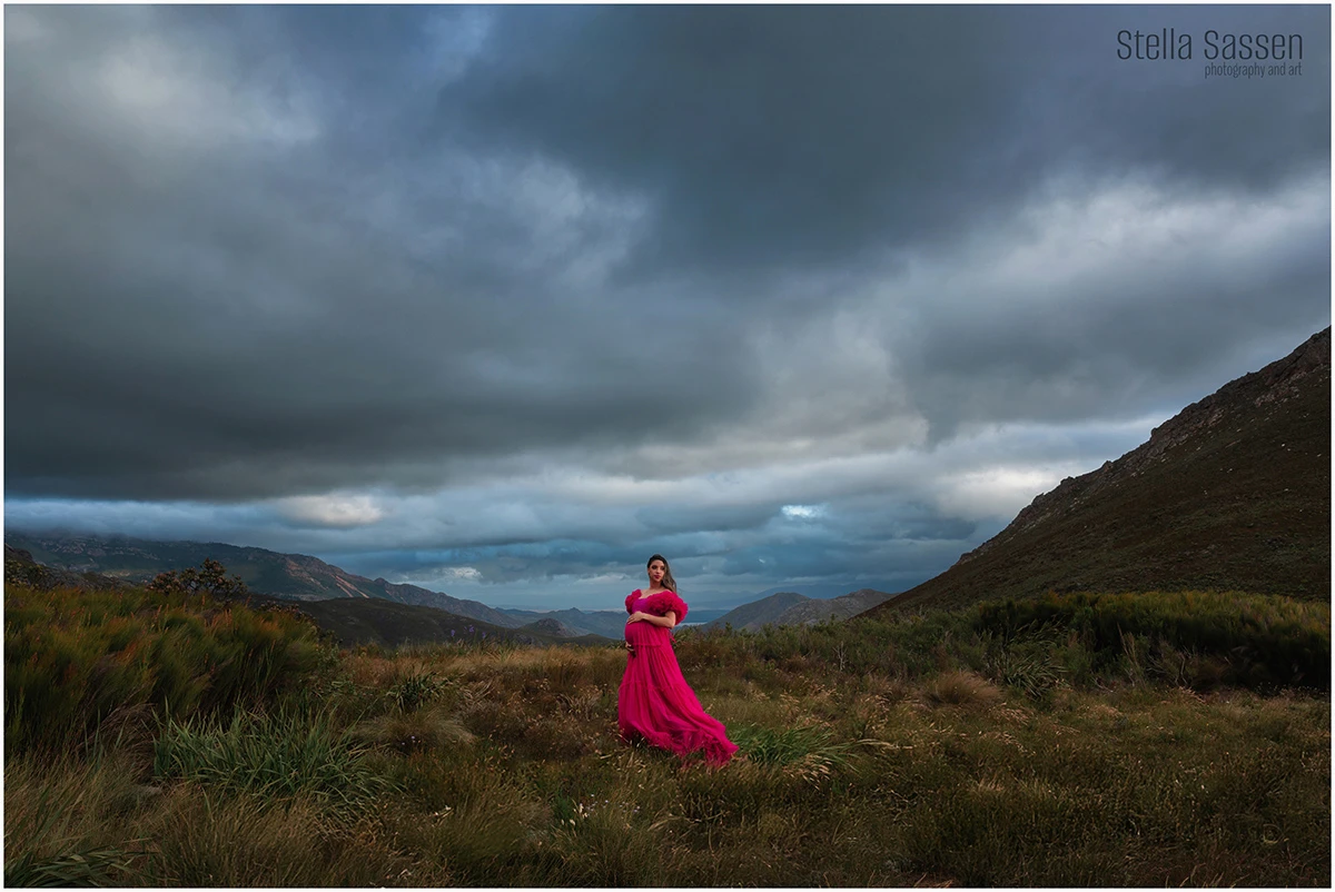  pregnant woman in a flowing hot pink gown standing alone in a dramatic mountain landscape near Franschhoek, dark storm clouds filling the sky behind her.